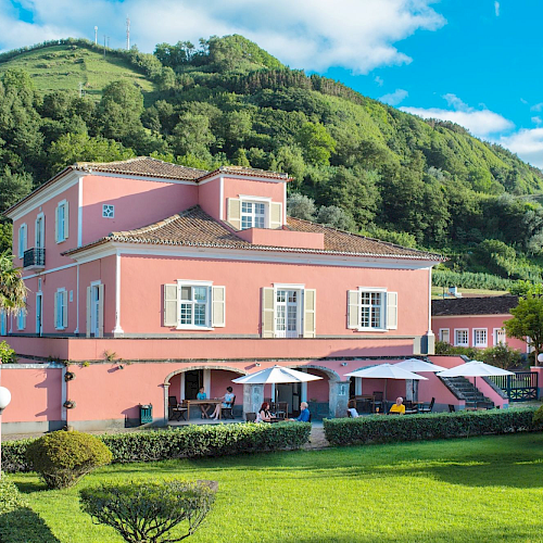 A pastel pink building with white trim sits in a lush, green landscape with mountains, blue sky, and a manicured lawn in front.