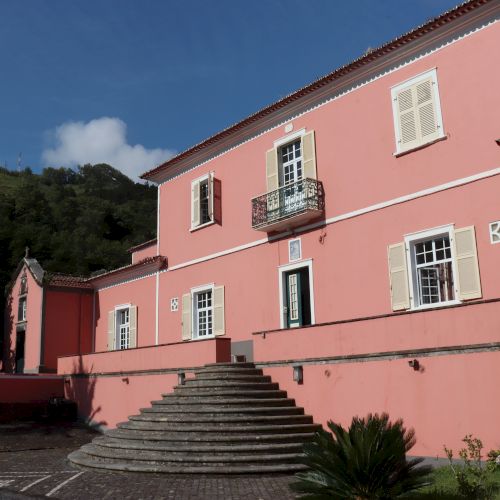 A large, pink, two-story building with multiple windows, green shutters, and a central staircase, surrounded by greenery and mountains in the background.