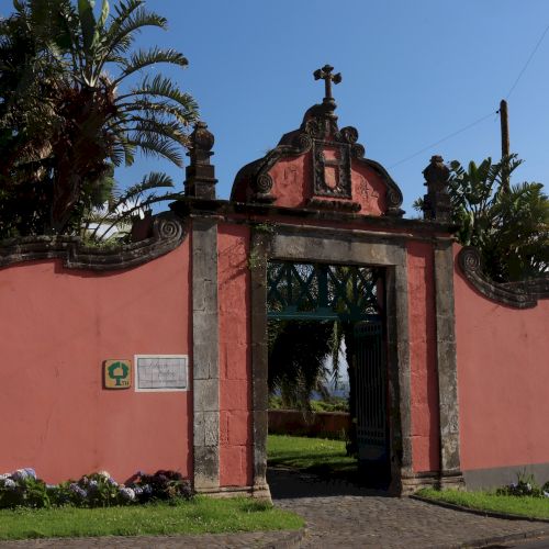 The image shows an ornate entrance gate with pink walls and palm trees, leading to a garden area, and a sign on the left side of the entrance.