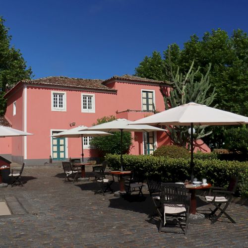 A pink building surrounded by greenery, with an outdoor dining area featuring tables, chairs, and large umbrellas on a paved courtyard, under a clear sky.